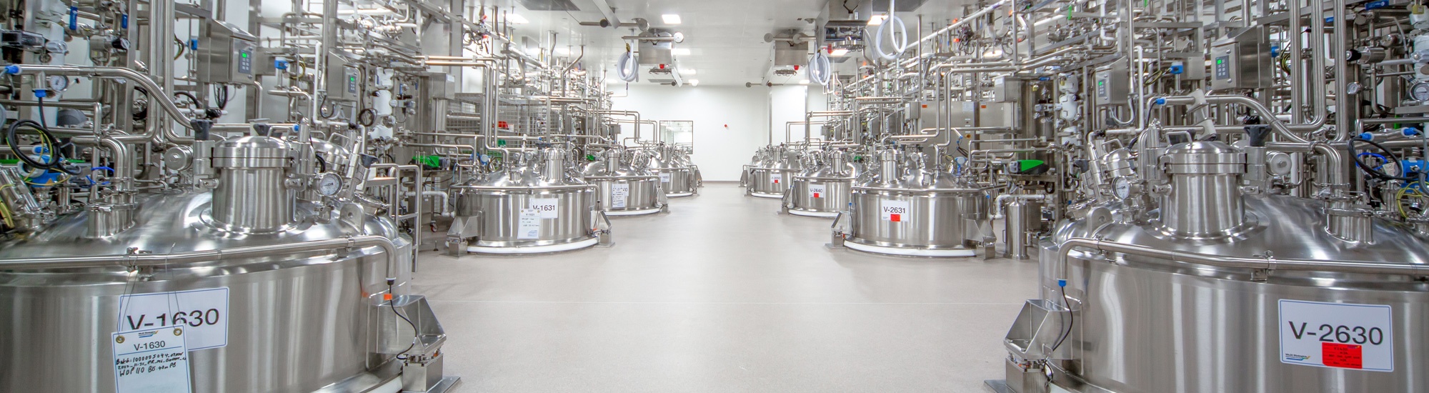 Salle de préparation des solutions tampons de WuXi Biologics. Deux files de cuves de production biologique en acier inoxydable sont présentées avec des tuyauteries de service en acier inoxydable, dans une chambre stérile très lumineuse.